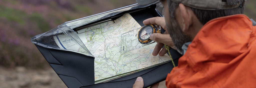 Person holding black clipboard containing map, with compass in right hand